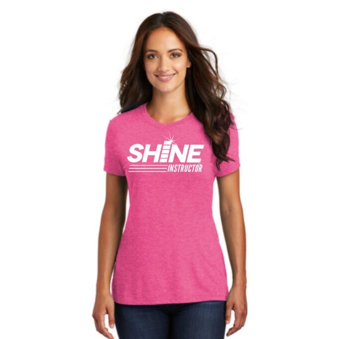 A woman wearing a bright pink T-shirt with the text "SHINE INSTRUCTOR" in white lettering stands facing forward, smiling.